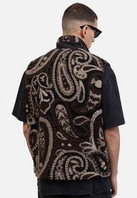 Rudas ir grietinėlės spalvos paisley rašto be rankovių liemenė su tekstūruotu fliso išoriniu paviršiumi, juoda apykakle ir nugaros detalėmis. Po ja vilkima juoda marškinėliai.