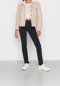 Person wearing beige corduroy jacket with white lining, cropped tie-dye shirt, black skinny jeans, and white chunky sneakers standing on gray floor.