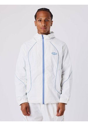 Giacca leggera bianca con zip e cuciture blu. Presenta un cappuccio e polsini elastici, con un logo discreto sul petto.