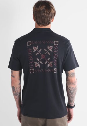 Chemise noire à manches courtes avec un grand motif floral violet et gris au dos, un logo discret et un col standard.