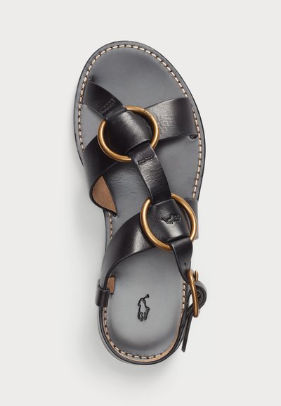 Melnas ādas sandales ar krustveida siksnām, zelta toņu gredzeniem un mīkstinātu iešuvumu. Iekļauj dekoratīvo audumu un regulējamu sprādzi.