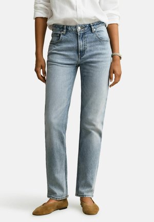 Persoon die lichtblauwe hoge taille rechte jeans draagt, een wit shirt met opgerolde mouwen, bruine platte schoenen en een gouden polshorloge.