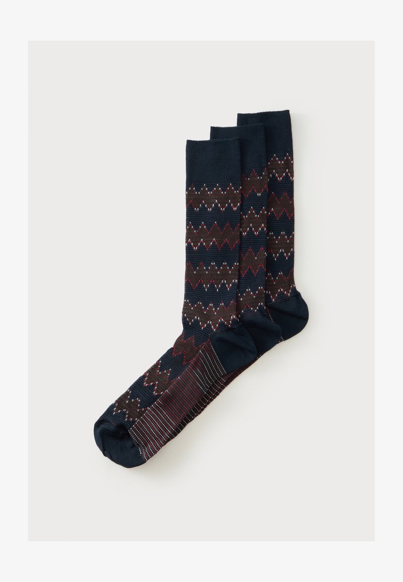 Trois chaussettes bleu marine avec des motifs en zigzag marron, rouge et blanc et des semelles rayées, posées à plat et superposées sur un fond blanc.