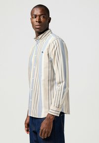 Camisa de manga larga a rayas en beige y azul claro, con cuello abotonado y un sutil acento con logo en el pecho; tela de algodón con una textura suave.