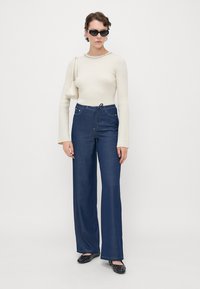 Marine brede ben-jeans med høy midje, med lommer og søm detaljer. Kombinert med en tettsittende elfenbensfarget ribbestrikket genser med lange ermer.