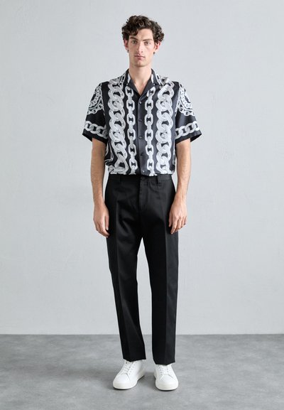 Short-sleeve black shirt with white chain pattern, paired with black trousers and white sneakers. Smooth fabric, relaxed fit, button-down front.