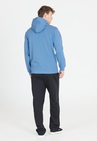 Blå huvtröja med långa ärmar, ribbad nederkant och mudd, i kombination med svarta sweatpants och svarta skor, sedd bakifrån.