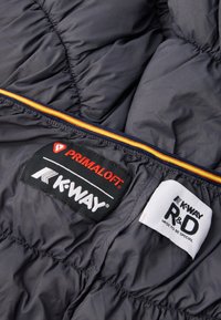 Εσωτερικό γκρίζας φουσκωτής ζακέτας με ετικέτες Primaloft και K-Way. Εντυπωσιακή πορτοκαλί και κίτρινη ριγέ λεπτομέρεια κατά μήκος του φερμουάρ. Απαλή υφή.