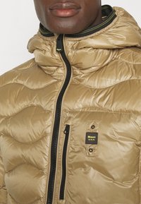 Giacca imbottita beige con cappuccio, dotata di chiusura con cerniera e tessuto strutturato, mostrata con una patch logo sul petto. Accenti neri visibili.