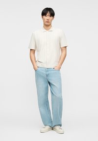 Νεαρός άνδρας που στέκεται με τα χέρια στις τσέπες, φορώντας κοντομάνικο κρεμ πουκάμισο, ανοιχτό μπλε τζιν και λευκά αθλητικά παπούτσια σε απλό φόντο.