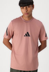 Gaiši rozā īsu piedurknu T-krekls ar apaļu kaklu. Priekšpusē ir melns Adidas logo un melnas akcenti uz piedurknēm. Gluda tekstūra.
