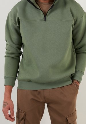 Mann trägt olivgrünes Sweatshirt mit Viertel-Reißverschluss und braune Cargo-Hose, Tätowierungen sichtbar an linker Hand und rechtem Handgelenk.