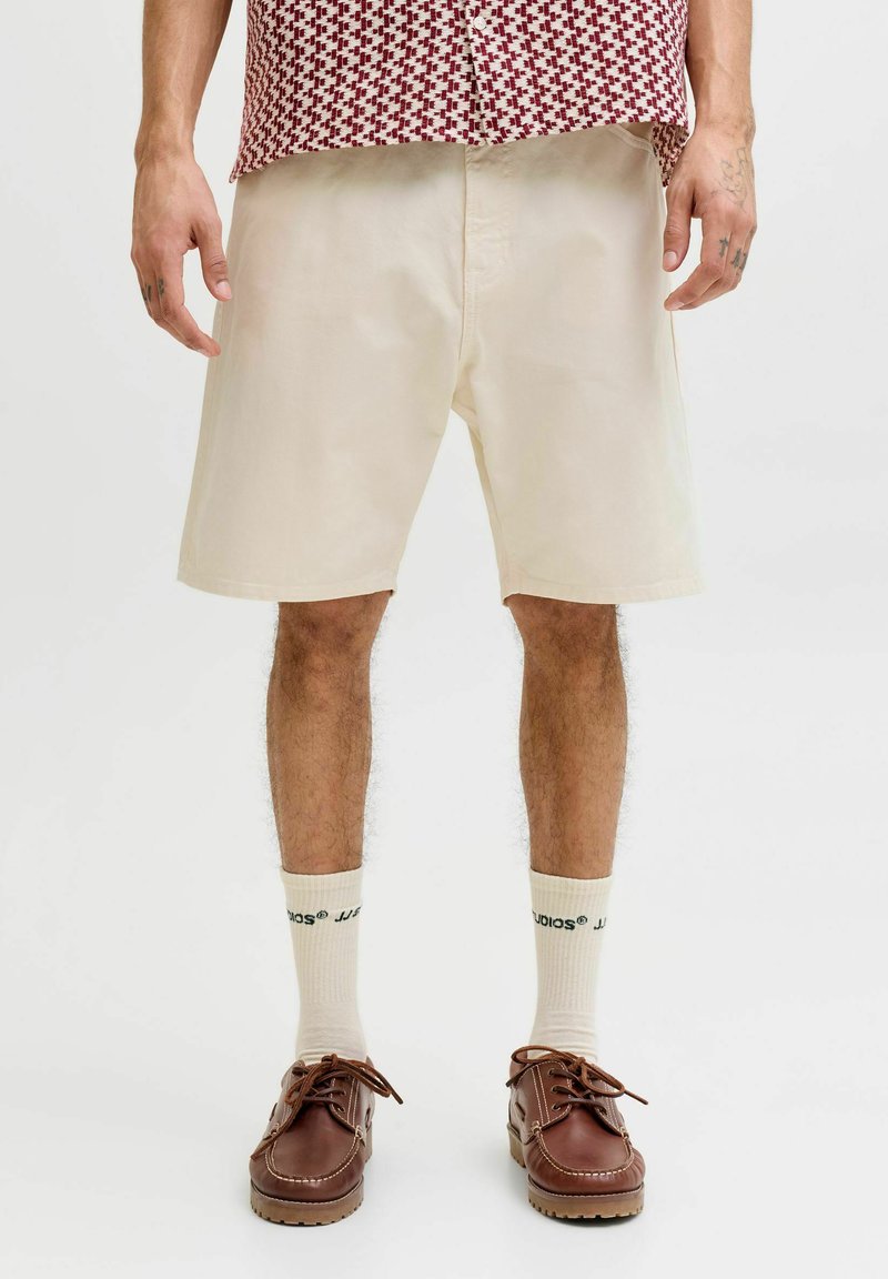 Homme portant un short crème mi-long, des chaussettes crème avec un texte noir, des chaussures en cuir marron et une chemise rouge et blanche à motifs.