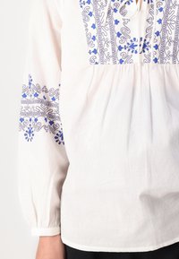Blusa bianca con ricami blu intricati sul colletto e sulle maniche, caratterizzata da una vita arricciata e maniche lunghe a sbuffo.