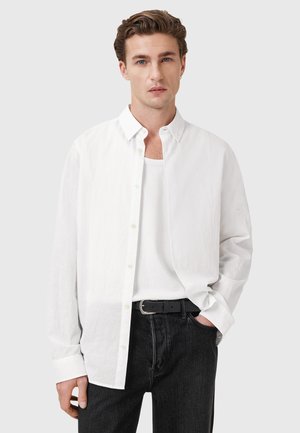 Camicia bianca a maniche lunghe in tessuto leggero, indossata aperta sopra una canotta bianca, abbinata a jeans scuri e una cintura nera.