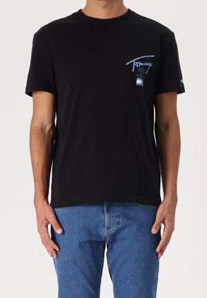 Un homme portant un t-shirt noir à manches courtes avec un texte graphique bleu sur la poitrine et un jean bleu, debout contre un fond uni.