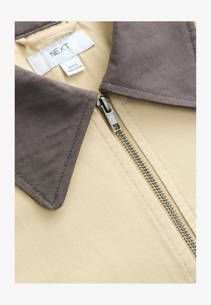 Tan takki harma harmaalta, jossa on metallinen vetoketju ja teksturoitu kangas. Tunnuslukuna "NEXT" ja "Valmistettu Bangladeshissa."