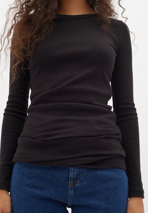 Person iført en tætsiddende sort langærmet skjorte og blå denimjeans, vist fra nakke til hofter mod en ensartet baggrund.