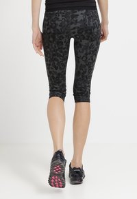 Persona con leggings a vita alta in leopardato nero e grigio e scarpe da corsa nere con dettagli rosa, in piedi su uno sfondo bianco.