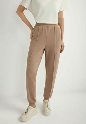 Kobieta w beżowych, zwężanych spodniach z bocznymi szwami, białej bluzce z krótkim rękawem i białych, sznurowanych trampkach, stojąca na jednolitym tle.