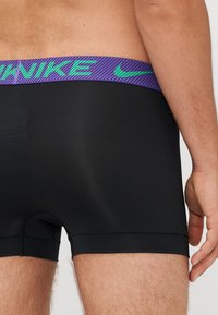 Črne atletske kratke hlače s tesnim krojem, ki imajo teksturiran vijolični pas z zelenim logotipom "Nike". Gladek material in šivi so zašiti.