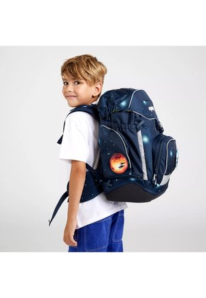 Young boy with light brown hair wearing a white shirt and blue pants carries a large navy blue space-themed backpack with planet patches.