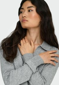 Hellgraues Langarm-Polohemd mit gerippten Bündchen, Kragen und weicher Textur. Das Modell posiert mit den Händen am Hals und an der Schulter.