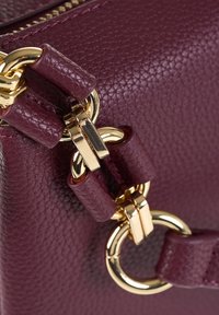 Primo piano di una tracolla in pelle bordeaux con texture, dettagli in anelli e ganci metallici dorati.