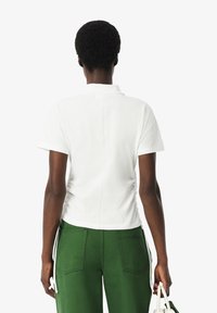 Witte polo met korte mouwen, voorzien van een kraag en geplooide zijnaad, gedragen met groene broek. Zachte katoenstructuur.