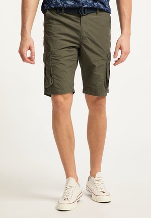 Hombre con pantalones cortos cargo verde oliva con bolsillos con solapa, cinturón azul marino, zapatillas blancas, de pie frente a un fondo claro y liso.