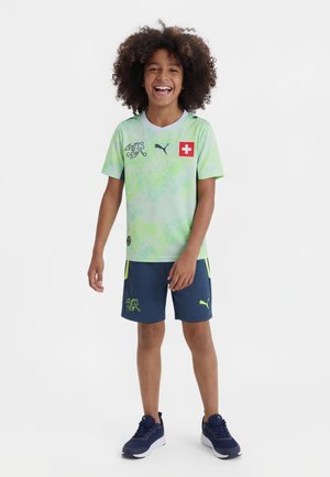 Nasmejan otrok s kodrastimi lasmi, oblečen v zeleno švicarsko nogometno majico, temno modre kratke hlače in temno modre superge, stoji na belem ozadju.