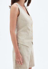 Beige linnelinneväst med V-ringning, ärmlös design, fem knappar framtill och sidofickor, kombinerad med matchande shorts.