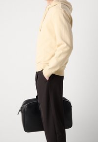 Sudadera beige con capucha y corte relajado, combinada con pantalones oscuros ajustados. Una bolsa de cuero negra está sostenida en la mano derecha del sujeto.