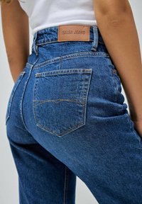 Pantalones vaqueros de mezclilla azul con una cintura alta, que presentan una etiqueta de cuero marcada "Salsa Jeans" y bolsillos traseros con detalles de costura en naranja.