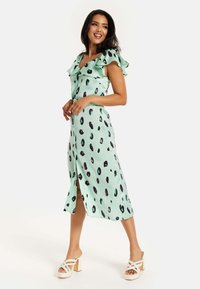 Mint green midi dress with a ruffled neckline and short sleeves, featuring black and purple spotted patterns. Side slit detail and smooth texture.