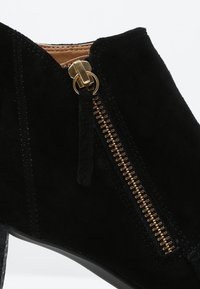 Bottines en suède noir avec fermeture éclair dorée et languette de tirage, présentant des coutures détaillées et un petit talon.