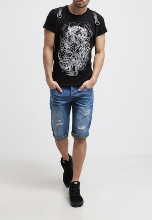 Camiseta de algodón negra con diseño gráfico en blanco, combinada con shorts de mezclilla claros y desgastados, con puños doblados, y zapatillas negras.