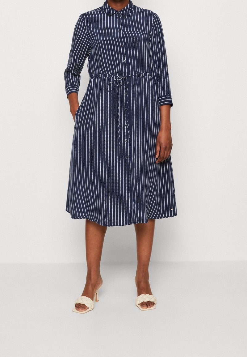 Femme portant une robe chemise midi bleu marine à rayures blanches verticales avec ceinture à la taille, associée à des sandales blanches à talons.