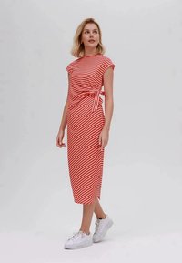 Vestido midi listrado em vermelho e branco com mangas curtas, detalhe de laço lateral e um corte descontraído, combinado com ténis brancos. Textura de tecido suave.