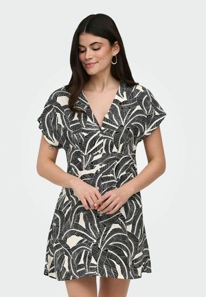Młoda kobieta w krótkiej sukience w czarno-biały wzór liści z krótkimi rękawami, stojąca ze splecionymi rękami i patrząca w dół.