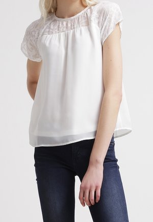 Femme portant un chemisier blanc à manches courtes avec des détails en dentelle aux épaules et un jean bleu foncé, debout devant un fond uni.