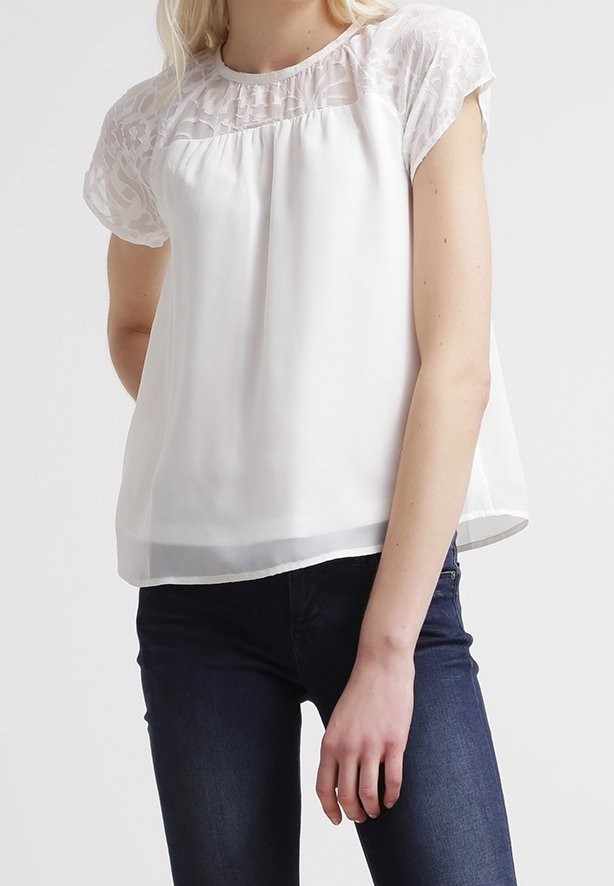 Femme portant un chemisier blanc à manches courtes avec des détails en dentelle aux épaules et un jean bleu foncé, debout devant un fond uni.