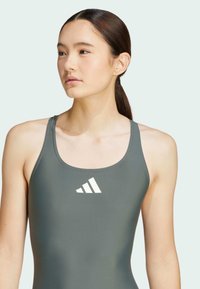Maillot de bain vert foncé avec une texture lisse et un design dos nageur, comportant un accent de logo blanc sur la poitrine. Style simple et fonctionnel.