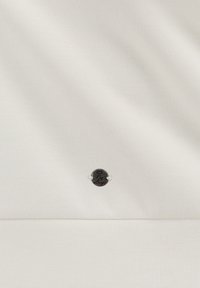 Vitt tyg med en mjuk textur har en liten svart logotypbricka med silverdetaljer, som visar texten "by Lory." Ren och minimalistisk design.