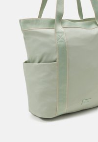 Sac fourre-tout vert clair en tissu durable, doté d'une poche latérale, de coutures contrastées et de poignées robustes. Texture lisse.