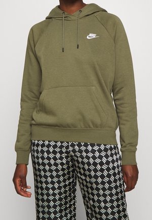 Person wearing olive green Nike hoodie with front pocket and black-and-white patterned pants, standing against a plain background.