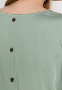 Hellgrüne Bluse mit glatter Textur, runder Ausschnitt und fünf dunkel gemusterten Knöpfen im Rücken.