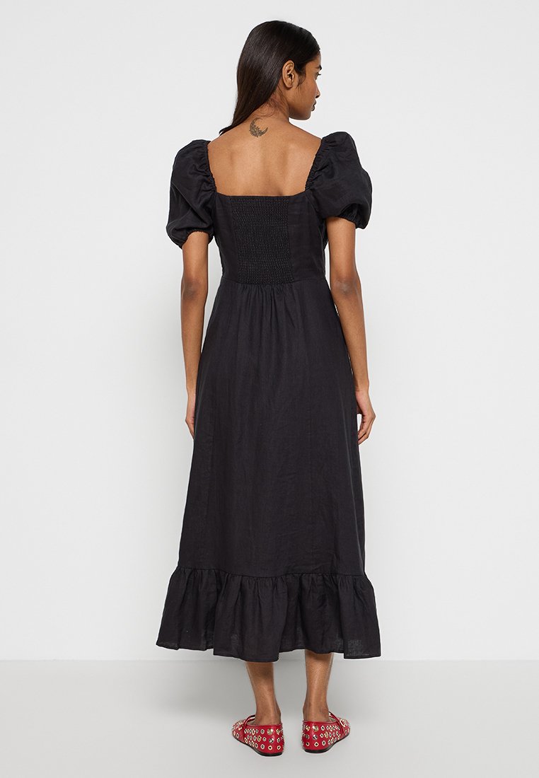 Robe maxi noire à manches bouffantes avec un dos smocké, un ourlet volanté et un tissu léger. Associée à des chaussures à enfiler à motifs rouges.