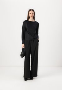 Top nero a maniche lunghe con scollo a barca, abbinato a pantaloni neri a gamba larga. La modella tiene una piccola borsa nera, in piedi su uno sfondo uniforme.