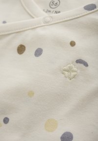 Weiche weiße Babykleidung mit Druckknöpfen am Hals, verziert mit verstreuten gelben, braunen und grauen Punkten sowie einem kleinen gestickten Design.